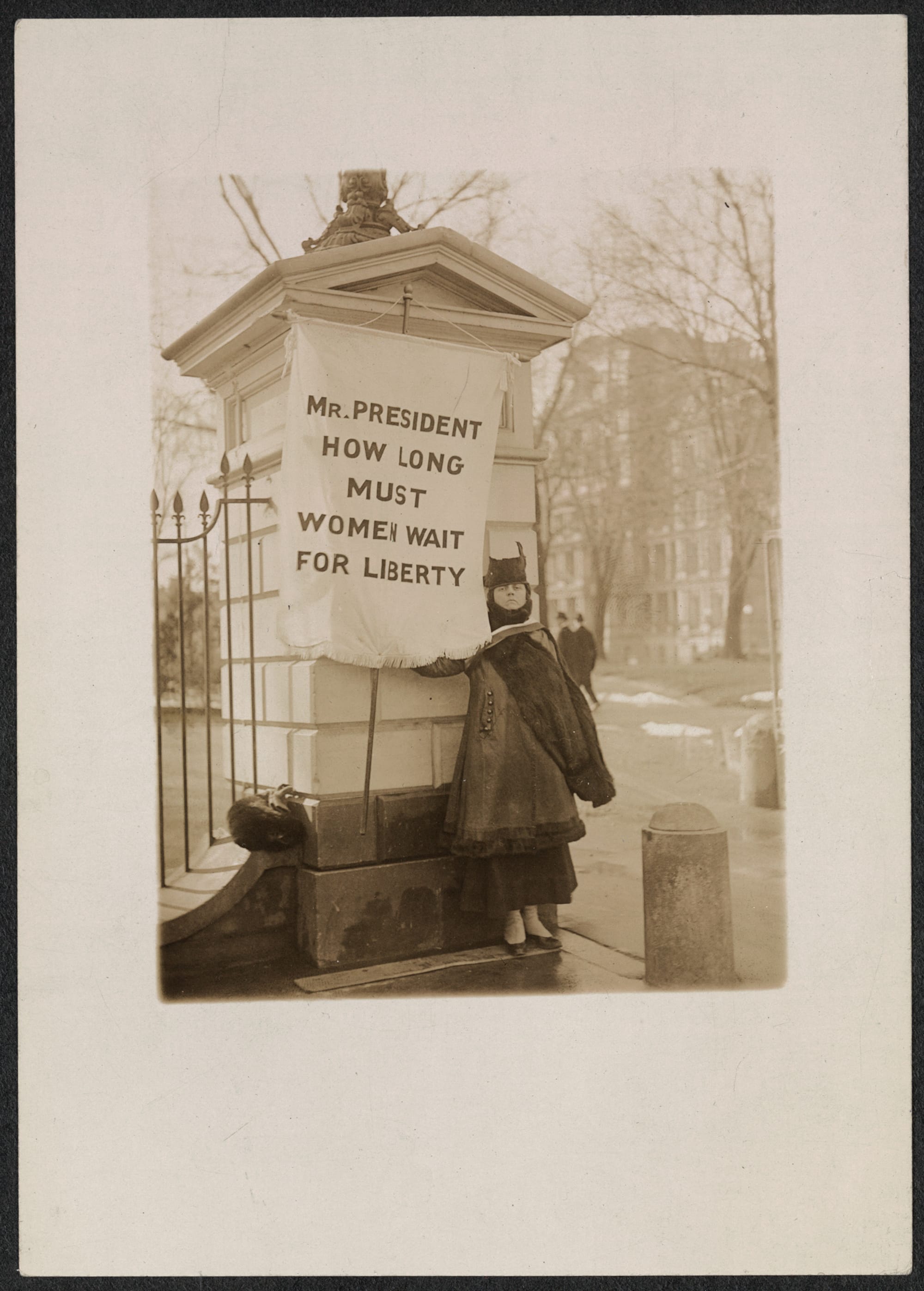 A sepia photo showing a woman holding a banner that reads: Mr. President, how long must women wait for liberty?