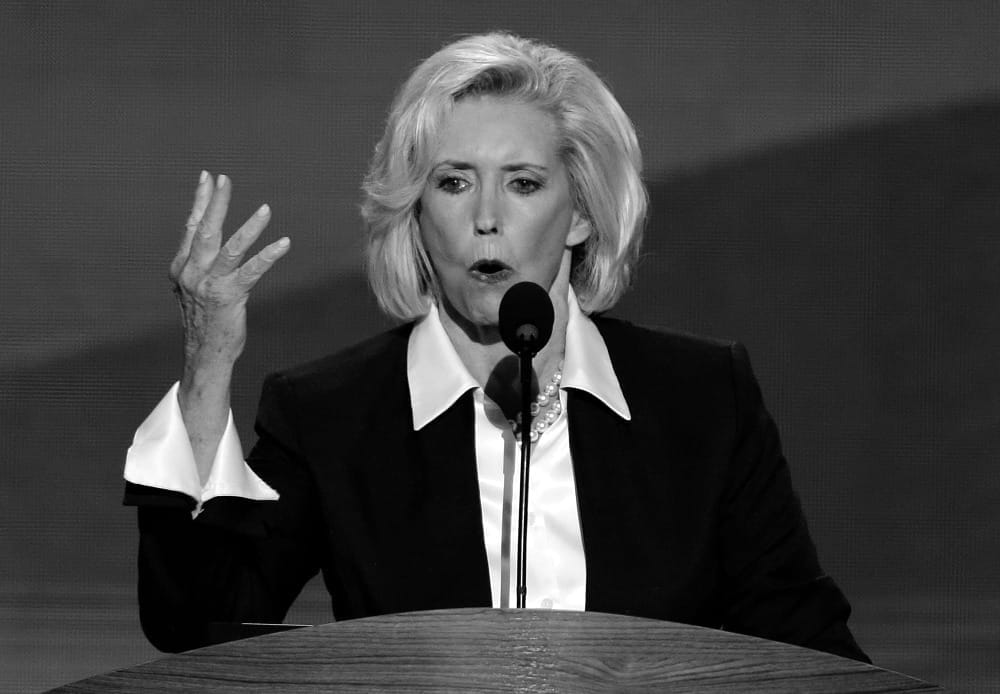 Black and white photo of Lilly Ledbetter at a podium. She has a blonde bob and wears a white shirt and black jacket