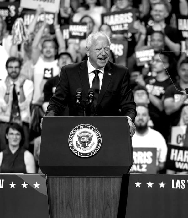 A black-and-white image of Tim Walz standing at a podium, with a crowd behind him.