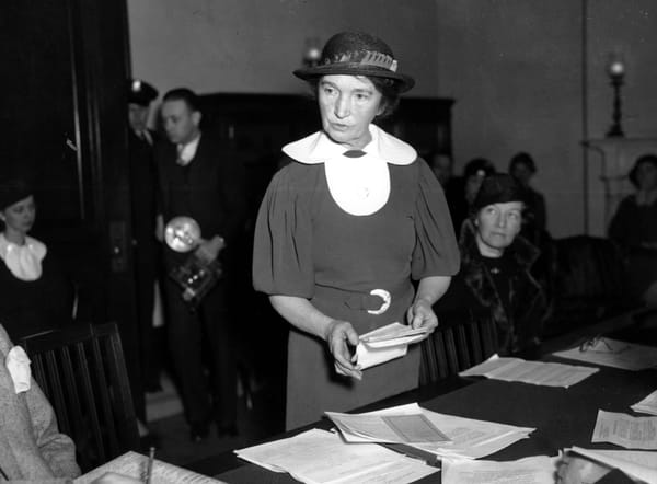 A black and white photo shows Margaret Sanger, wearing a hat and belted dress, standing at a table covered with documents.