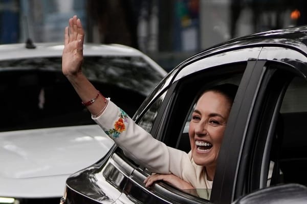 Claudia Sheinbaum waves from the window of a car. She is smiling broadly.