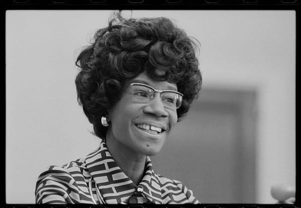 A black and white headshot of Shirley Chisholm. She wears glasses and a geometric print blouse. 