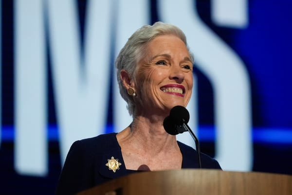 Cecile Richards smiles widely at a lectern. She wears navy, with gold earrings, a gold broach and cerise lipstick.